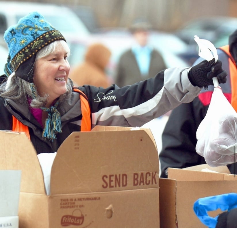 Smiling older woman, bundled in a winter coat and stocking hat, stands in front of a conveyor belt filled with boxes. She holds out a bag of food, surrounded by a snowy outdoor setting.
